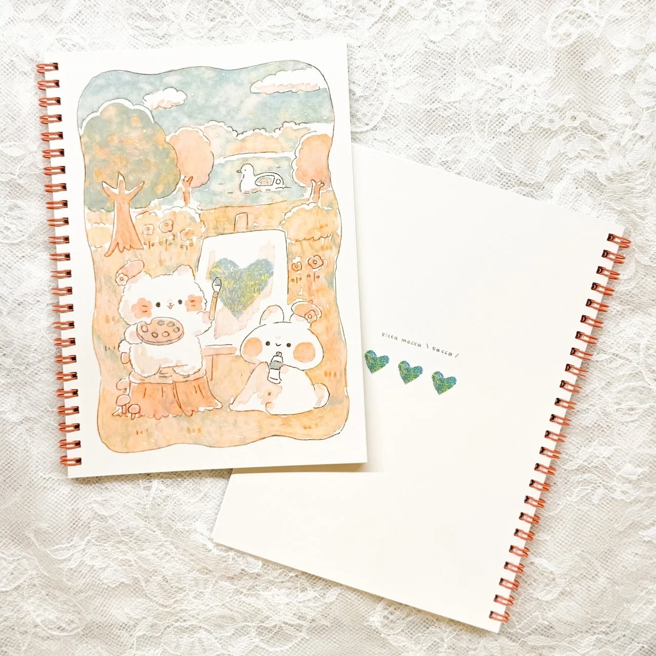 白いレースの布の上に置かれた2冊のリングノート。1冊は動物たちが絵を描く可愛らしい水彩イラスト