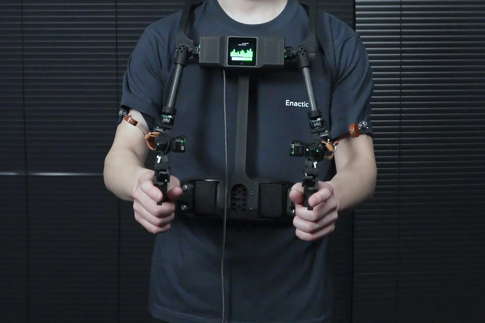 男性が胸元に装着型のデバイスを身につけ、両手にコントローラーを握っている様子。デバイスにはディスプレイがあり、Tシャツには「Enactic」の文字が見える。