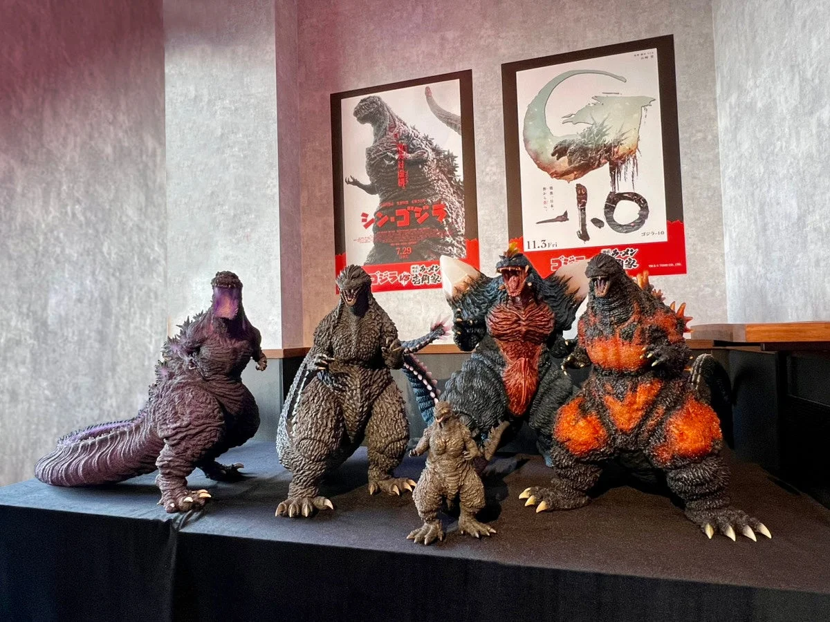 「シン・ゴジラ」や「ゴジラ-1.0」などの映画ポスターを背景に、様々な形態のゴジラフィギュアが展示されている様子