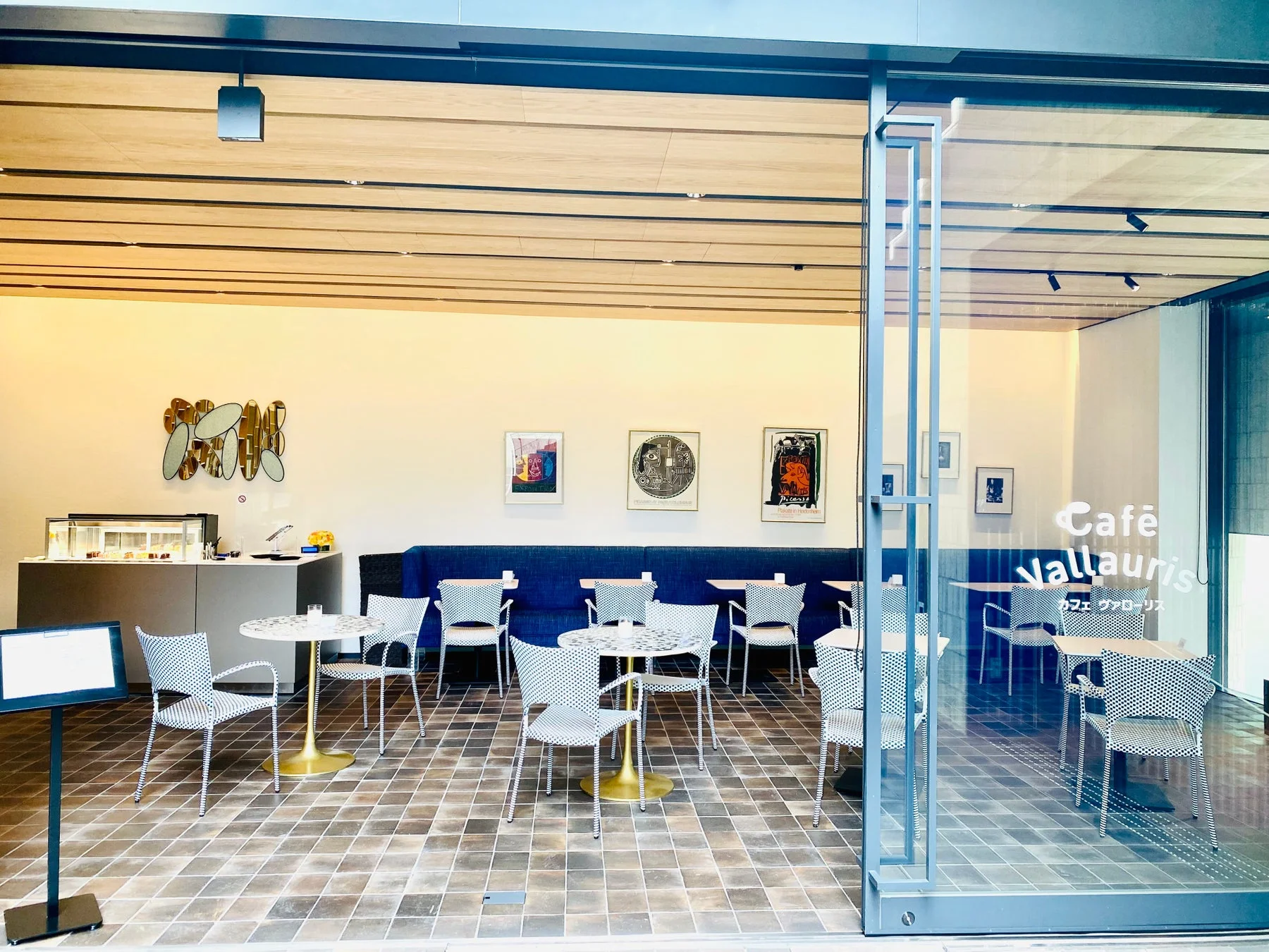 美術館のような空間に設置されたモダンなカフェの入口の様子。ドット柄の椅子と白いテーブルが並び、壁にはアート作品が飾られています。店内にはデザートケースも見え、落ち着いた雰囲気でくつろげそうです。