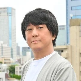 都会のビル群を背景に、黒髪のアジア人男性が白いTシャツとグレーのカーディガンを着用し、カメラを見つめているポートレート写真です。穏やかな表情で、カジュアルな印象を与えます。