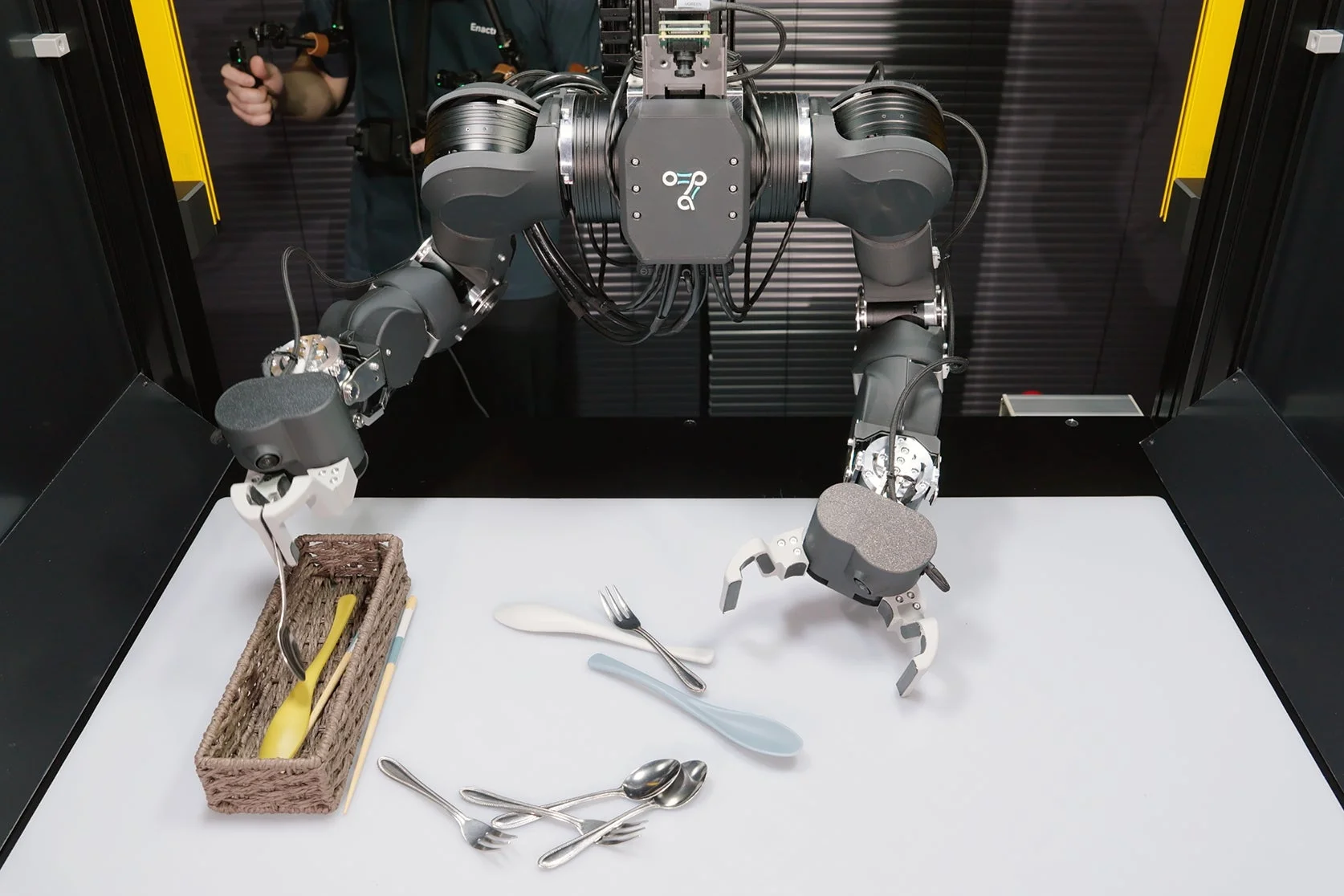 ロボットアームがテーブル上のカトラリーを操作している様子。一人の人物がロボットを遠隔操作しているように見え、ロボットの器用な作業を示している。