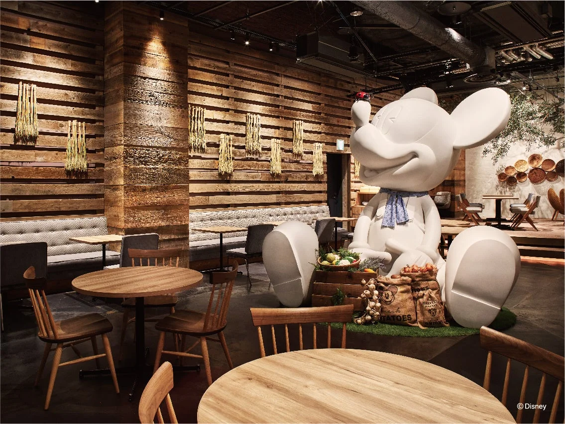 木目調の壁と温かみのあるインテリアが特徴のレストランまたはカフェの店内。中央には大きなミッキーマウスの像が座り、その周りには収穫されたばかりのような野菜やポテトが飾られ、農園風の素朴でおしゃれな空間を演出しています。