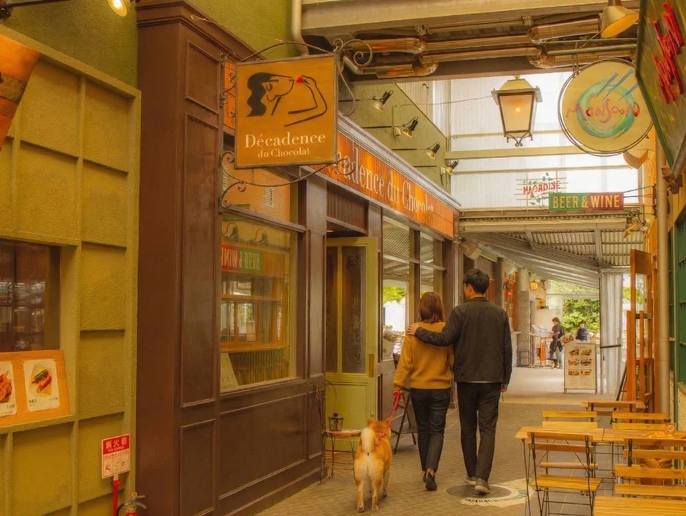 犬を連れたカップルが、レトロな雰囲気の商店街を歩いている様子を捉えた画像です。