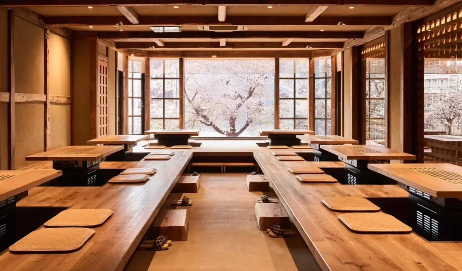 大きな窓から満開の桜の木が見える、和風の趣のあるレストランの内部です。木製のテーブルと掘りごたつ式の座席が整然と並び、落ち着いた雰囲気の中で食事が楽しめる空間が広がっています。