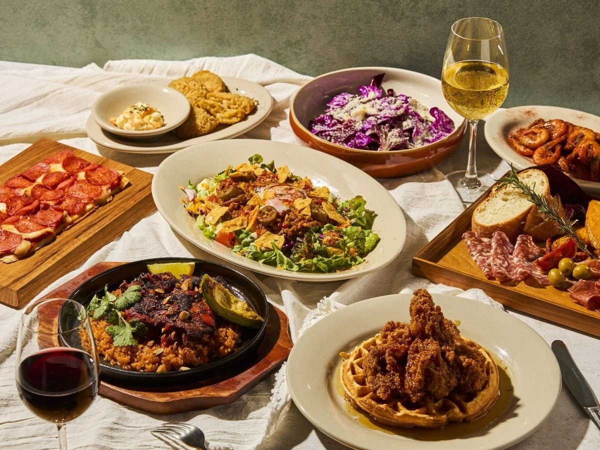 テーブルいっぱいに多様な料理が並べられた豪華な食事の風景。ペパロニピザ、フライドチキンとワッフル、サラダ、シャーキュトリーボード、そして赤白ワインが食欲をそそり、特別な日のごちそうやパーティーの賑やかな雰囲気を伝えています。