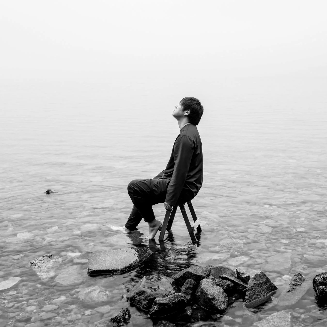 モノクロームの写真で、浅い水の中に置かれた椅子に座った人物が、霞んだ水平線を見上げている。ミニマルな水辺の風景の中で、孤独、思索、そして静寂の感覚を伝える一枚。