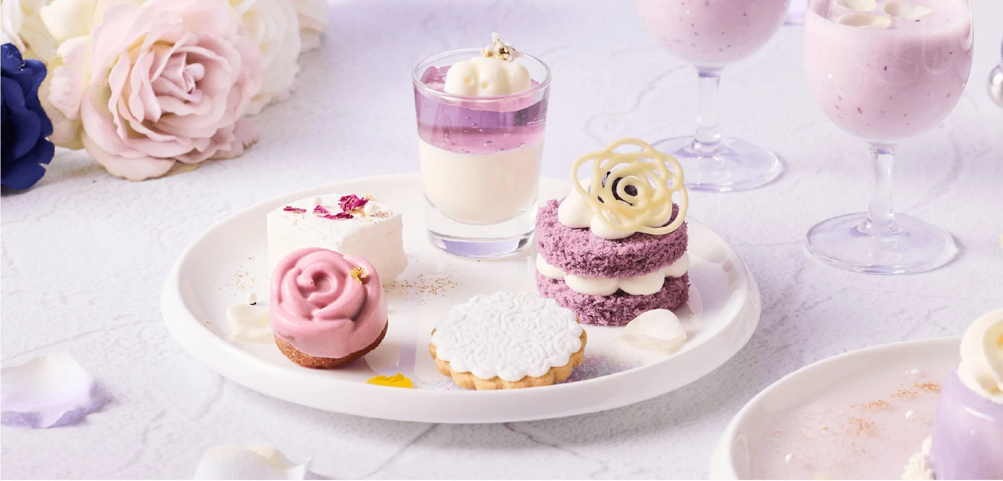 白を基調としたテーブルに、紫やピンクの華やかなデザートとドリンクが並べられた写真です。バラの形をしたムース、層になったケーキ、ゼリー、クッキーなど、上品で美しいスイーツがティータイムを彩ります。
