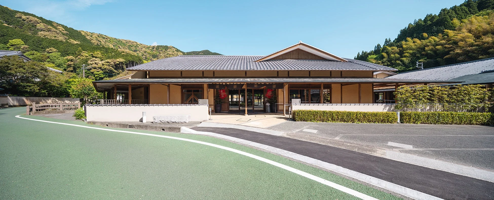 青い空と豊かな緑の山々に囲まれた、瓦屋根の伝統的な日本家屋の風景。建物前には道がカーブし、入り口には「壽」の文字が入った赤い飾りが印象的。