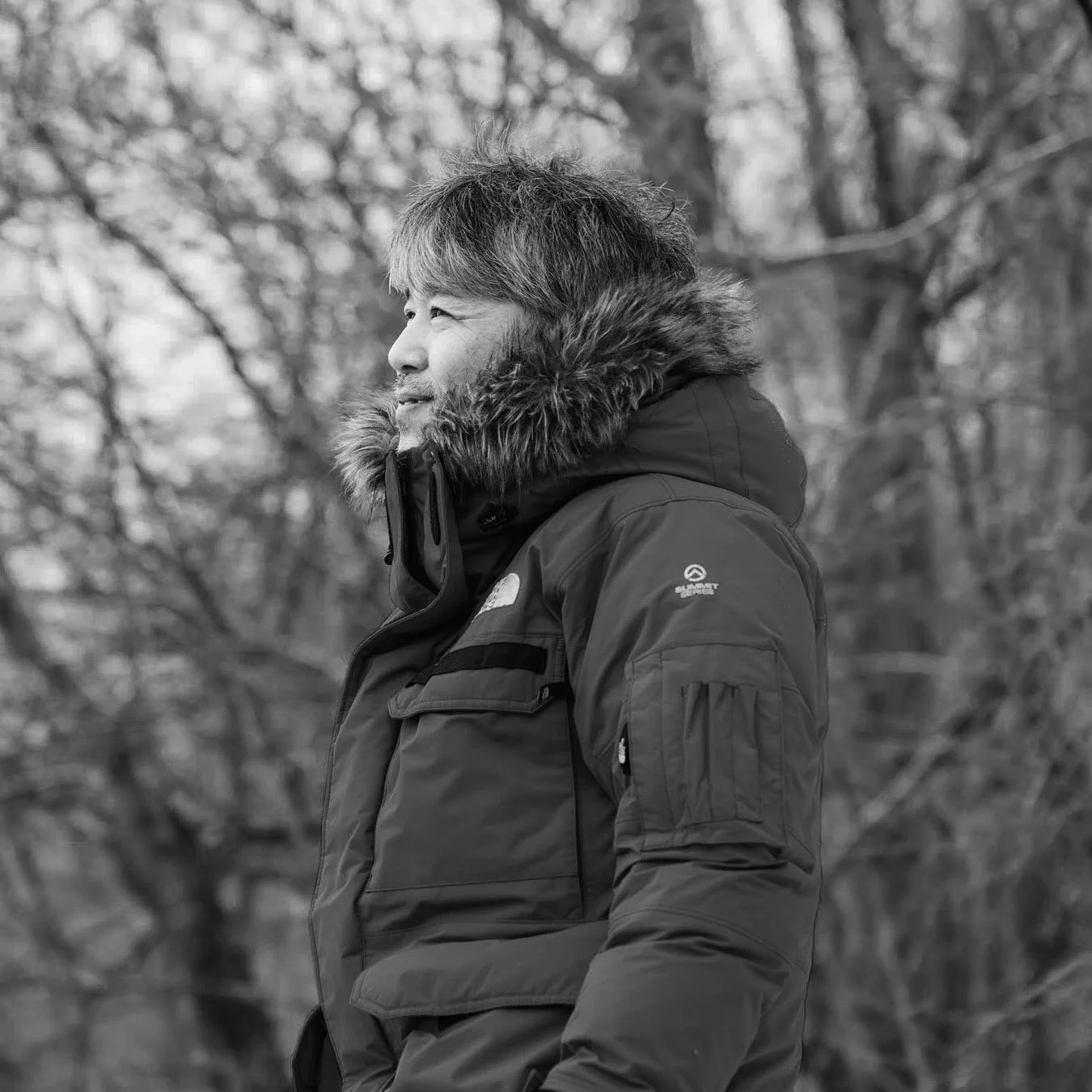 ファー付きの防寒着を着用した男性が、屋外で遠くを見つめているモノクロ写真です。