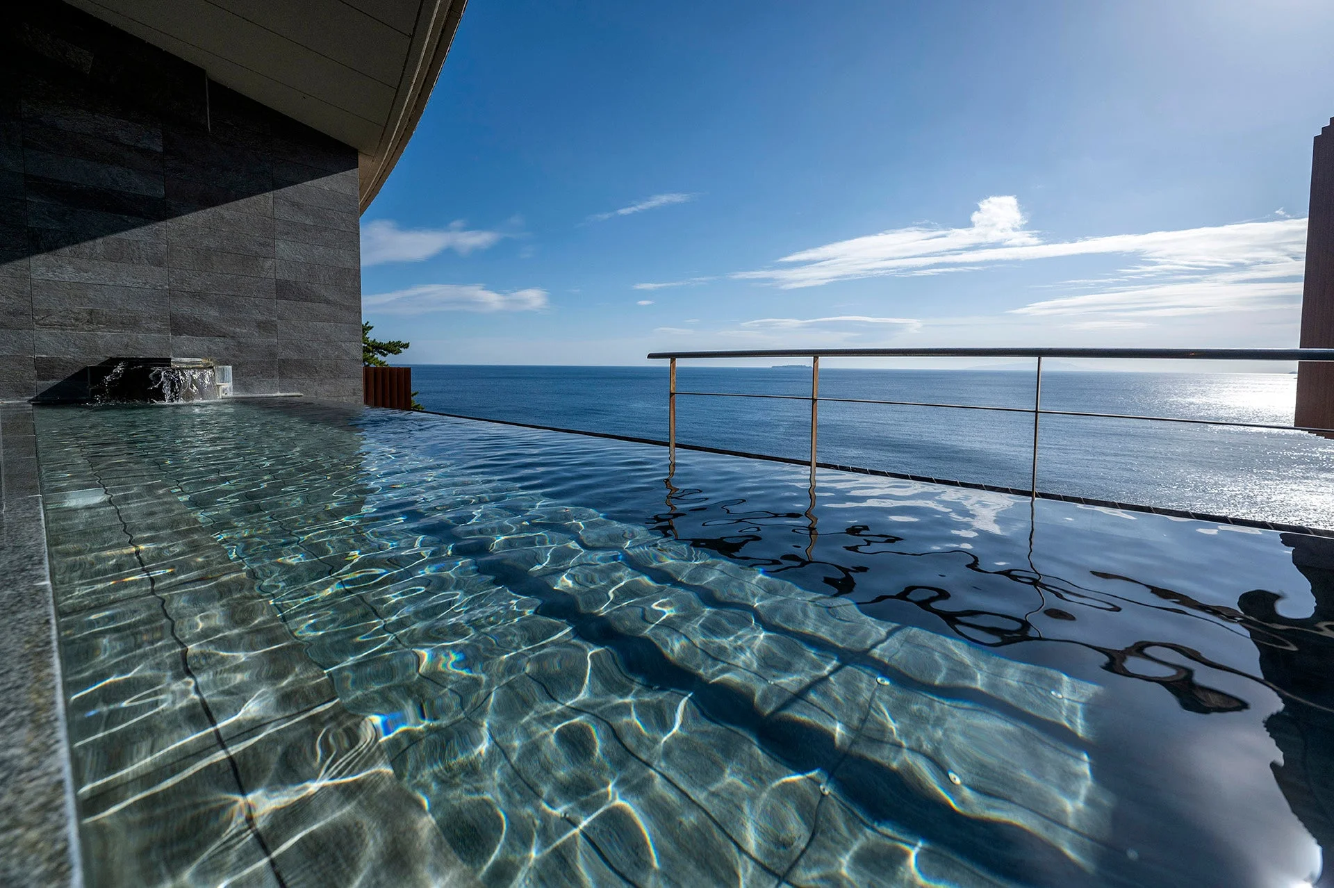 晴れた日の青空の下、広がる海を望むインフィニティプールまたは露天風呂の風景。水面に太陽の光が反射し、波紋が美しい。贅沢なリゾート気分を味わえる開放的な空間です。