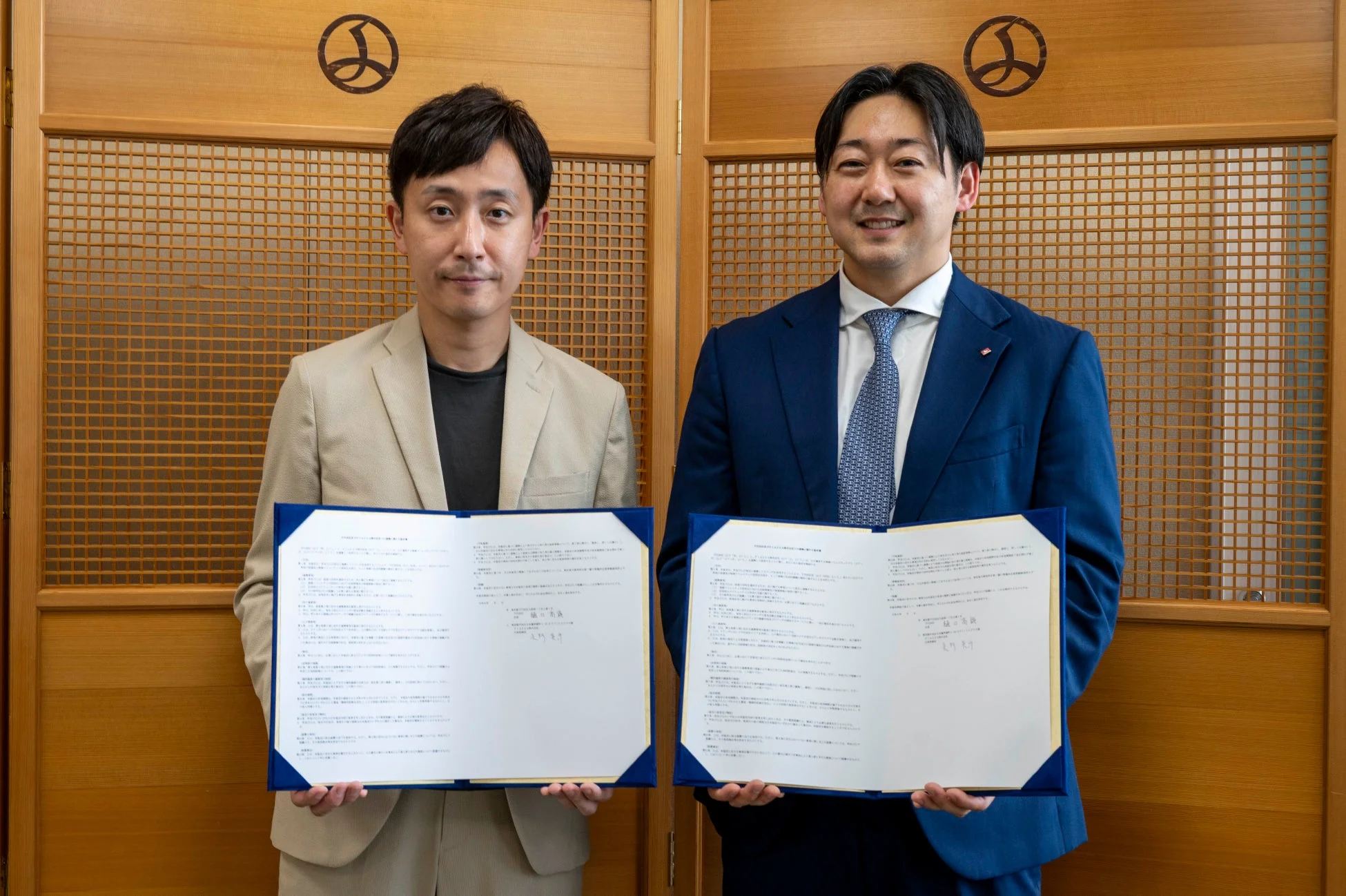 和風の背景の前で、二人の男性がそれぞれ協定書のような書類を手に持ち、笑顔で立っている