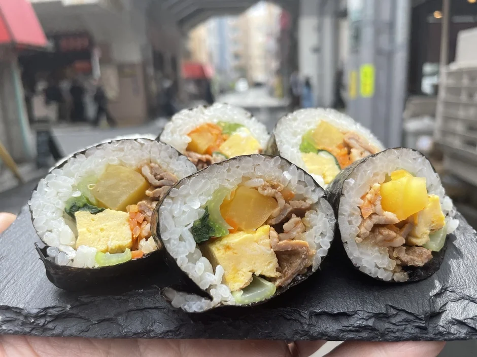 屋外で撮影された、スレート調の皿に盛り付けられたキンパのクローズアップ写真です。米、卵、肉、野菜が詰まった色鮮やかな海苔巻きで、美味しそうな一品です。