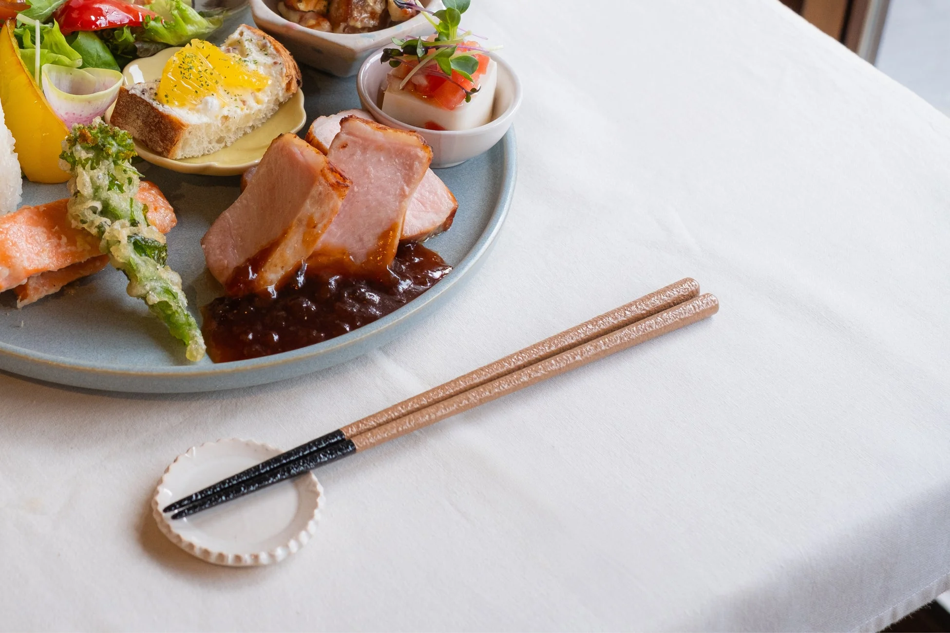 白いテーブルクロスに、肉料理、魚、野菜の天ぷら、卵パン、豆腐、サラダなど、彩り豊かでバランスの取れた和風プレート料理が美しく盛り付けられています。箸が添えられ、食欲をそそる一食の風景です。