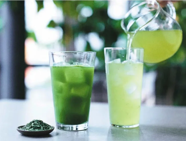 氷が入った2つのグラスに緑色の飲み物が注がれており、片方は濃い緑、もう片方は薄い緑です。ピッチャーから薄い緑の飲み物がグラスに注がれ、手前には乾燥した緑の葉が置かれています。