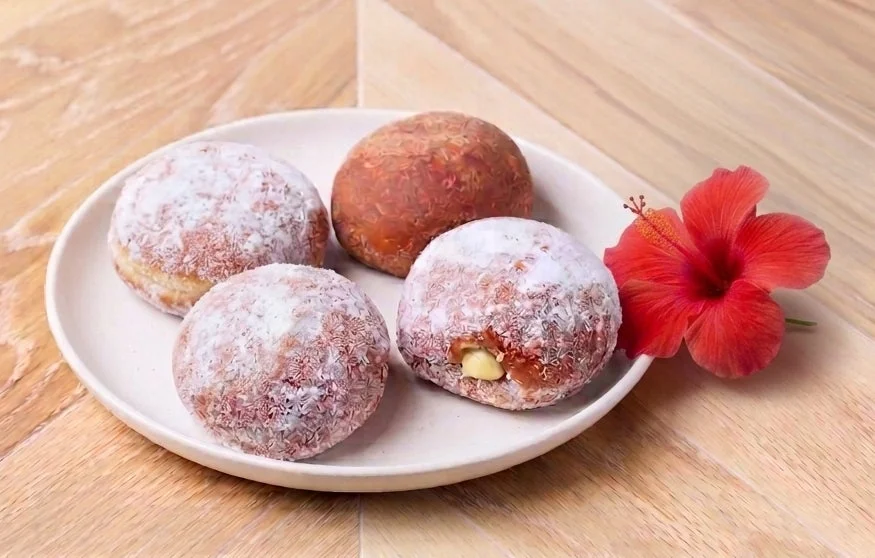 白いプレートに4つのドーナツまたはマラサダが盛られており、そのうち3つは粉砂糖で覆われています。1つからはクリームが溢れ出ています。プレートの横には鮮やかな赤いハイビスカスの花が添えられ、木目調のテーブルに置かれています。