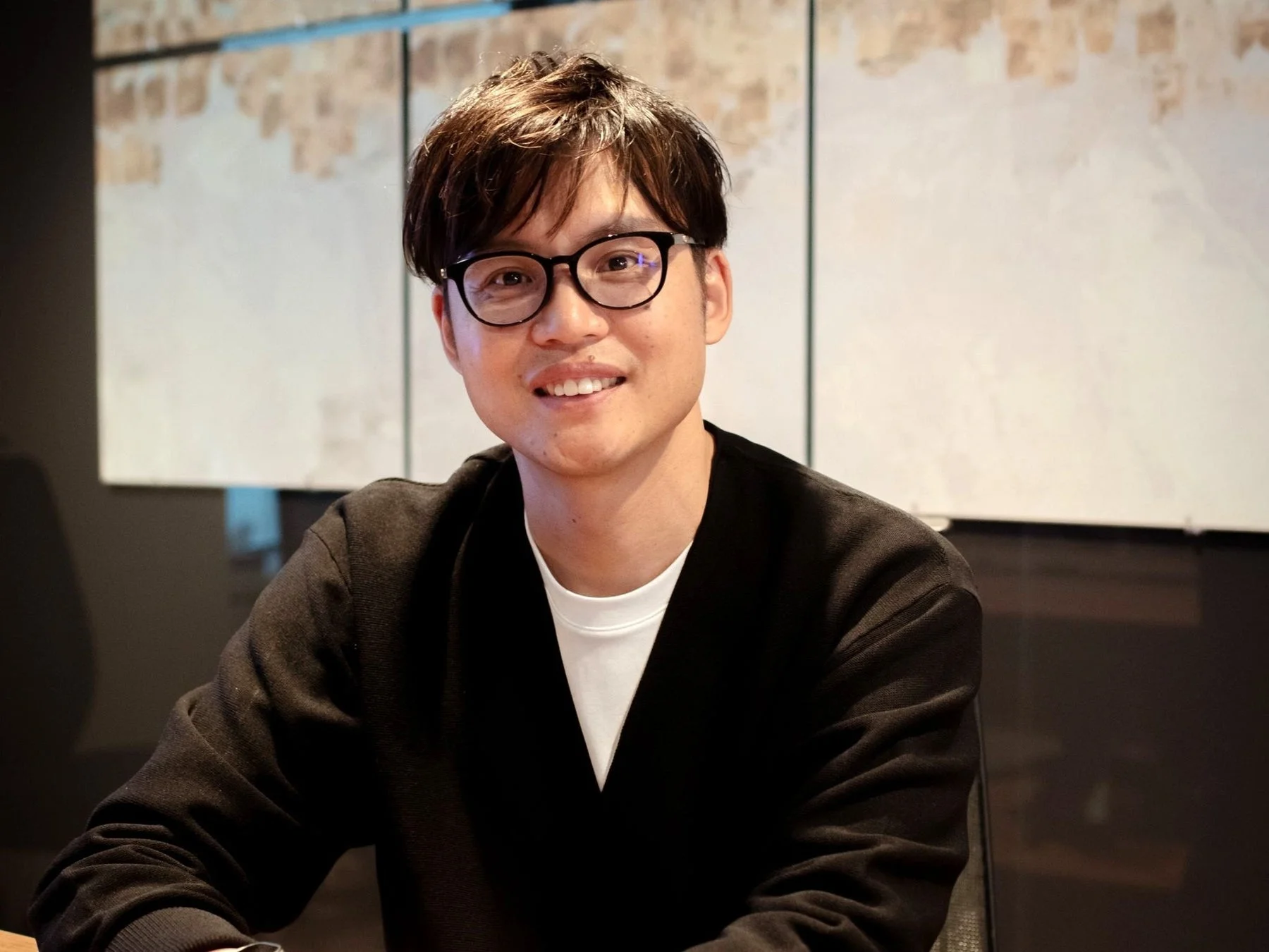 眼鏡をかけたアジア人男性が、黒いカーディガンと白いTシャツを着てにこやかに座っているポートレート。背景はぼやけている。