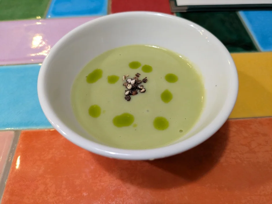 カラフルなタイル柄のテーブルに置かれた白い器に入った、緑色の冷製スープです。オリーブオイルのような緑色の液体と黒い粒状のスパイスがトッピングされています。