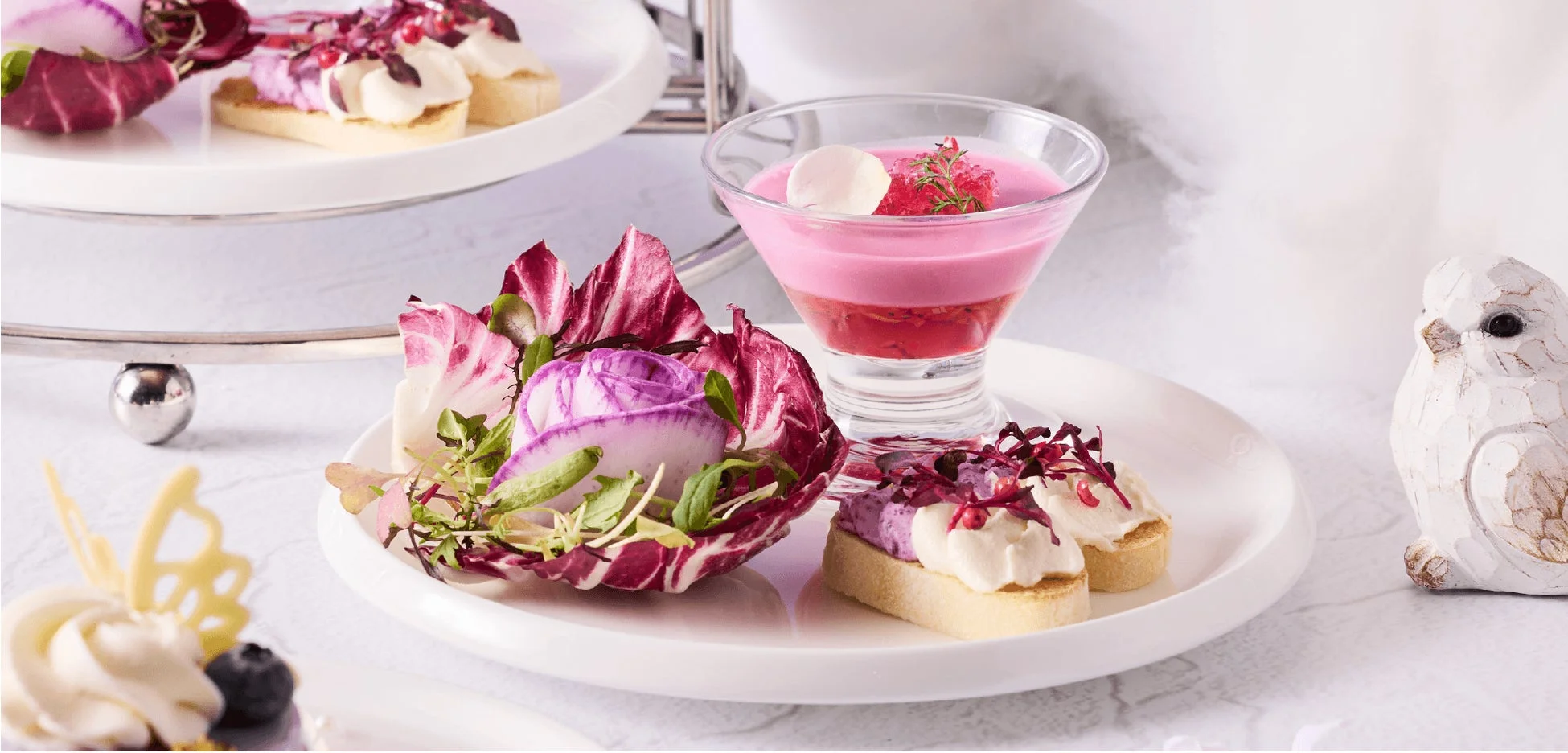 ピンクとパープルを基調とした華やかなアフタヌーンティーセット。層になったピンクのドリンク、クリームが乗ったカナッペ、ラディッシュでバラを模したような美しいサラダが並び、上品な雰囲気を演出しています。可愛らしい鳥の置物も添えられています。
