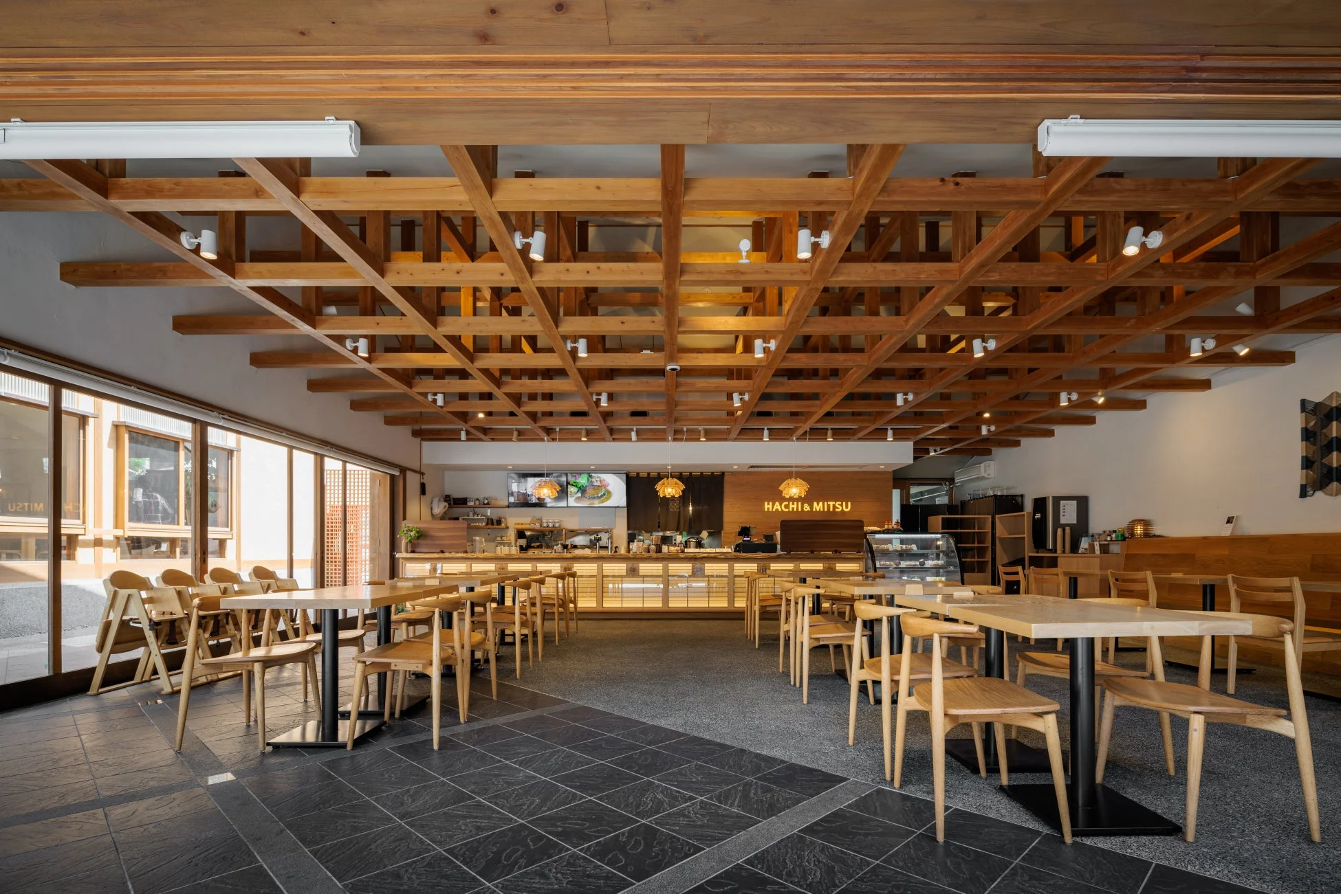 木材を多用したモダンで広々としたレストランまたはカフェの店内。天井の格子状の梁や木のテーブルと椅子が特徴的で、奥にはカウンターと「HACHI & MITSU」のロゴが見える。