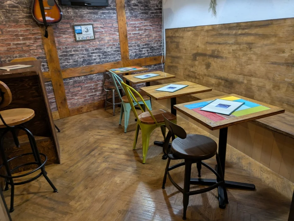 レンガ壁と木目調の壁が特徴的なカフェまたはバーの店内風景。木製のテーブルとカラフルな椅子が並び、壁にはギターが飾られている。ヘリンボーン柄の床がおしゃれな雰囲気を演出している。