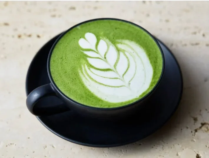 黒いカップに入った鮮やかな緑色の抹茶ラテで、美しいラテアートが施されています。ベージュ系の石のようなテーブルに置かれており、落ち着いた雰囲気です。