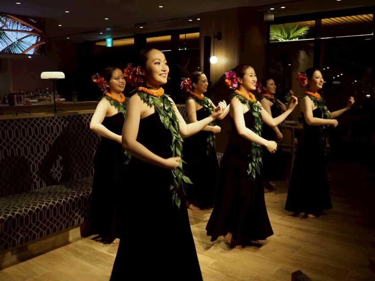 黒いドレスと鮮やかなレイを身につけた女性たちが、笑顔でフラダンスを踊っています。リゾートホテルのような場所で、優雅なハワイアンパフォーマンスを披露している様子です。