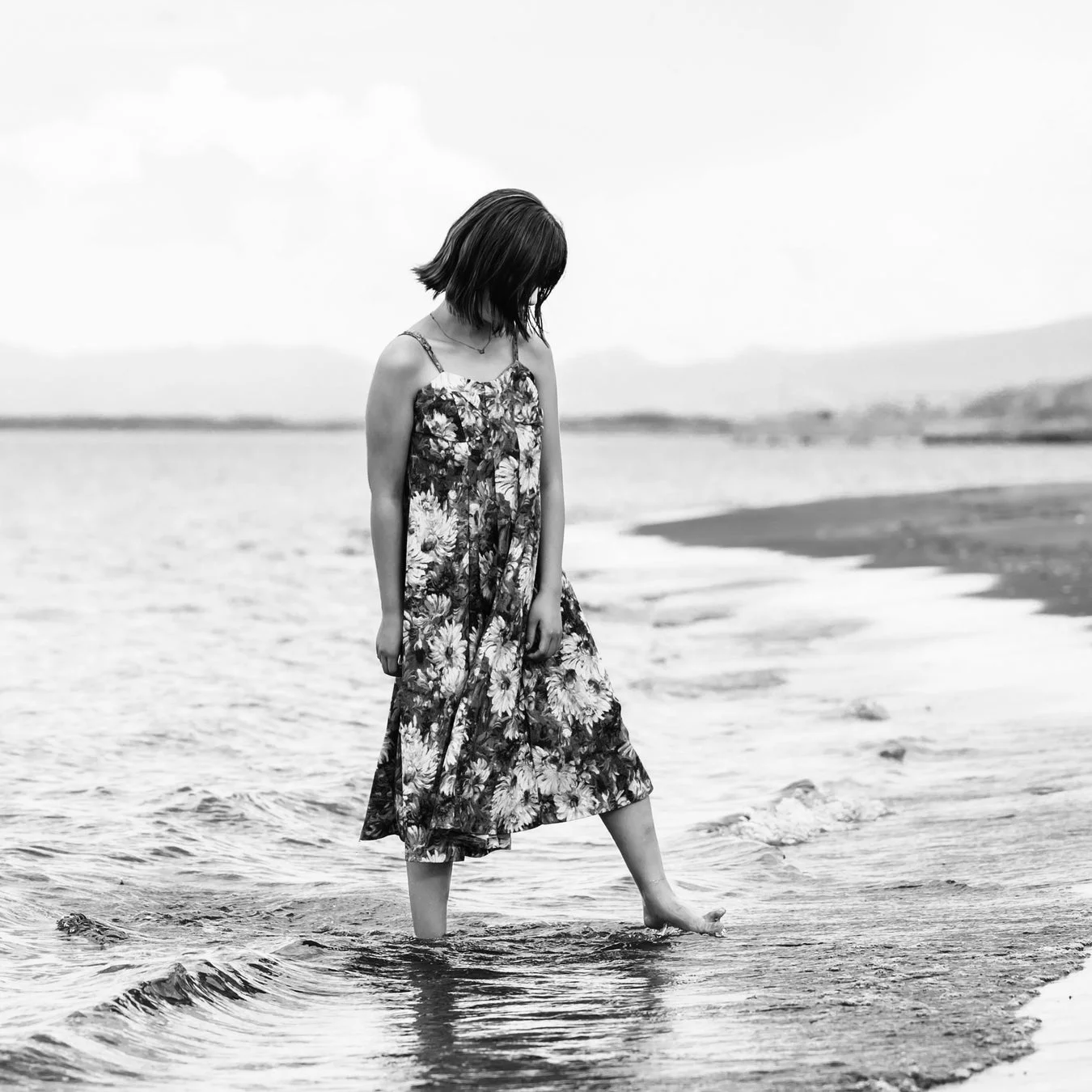 花柄のワンピースを着た女性が水辺に立ち、うつむいている姿を捉えたモノクロ写真です。水面に波紋が広がり、静かで思索的な雰囲気が漂っています。