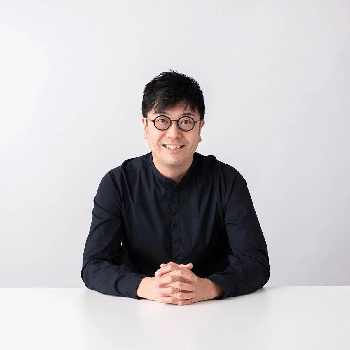 白背景の前で、黒いシャツと丸眼鏡をかけた男性がテーブルに座り、両手を組んで笑顔を見せているポートレート写真