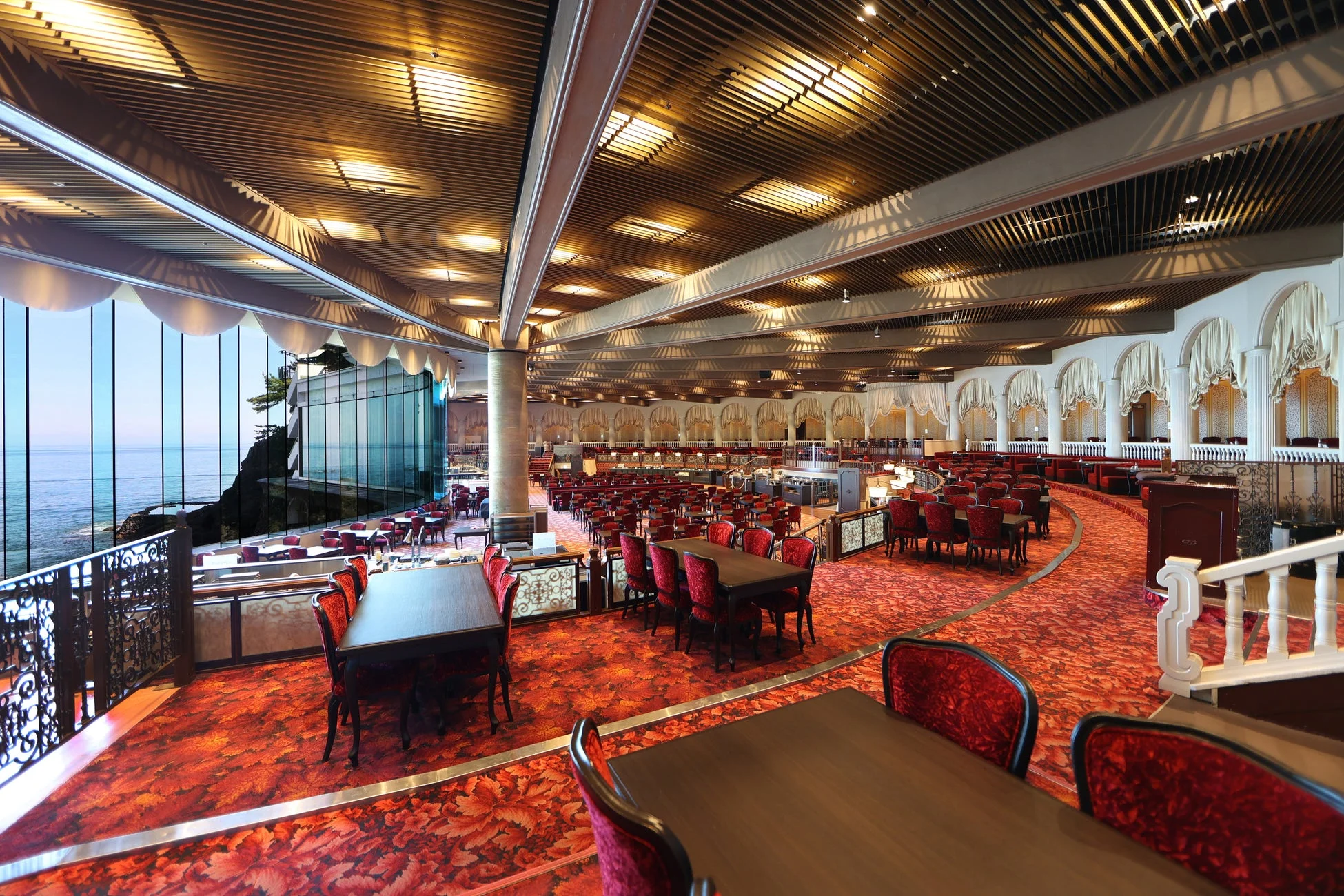 海を一望できる豪華なレストランまたは宴会場の広々とした内装。赤い絨毯とベルベットの椅子が特徴的で、優雅な雰囲気が漂う空間です。