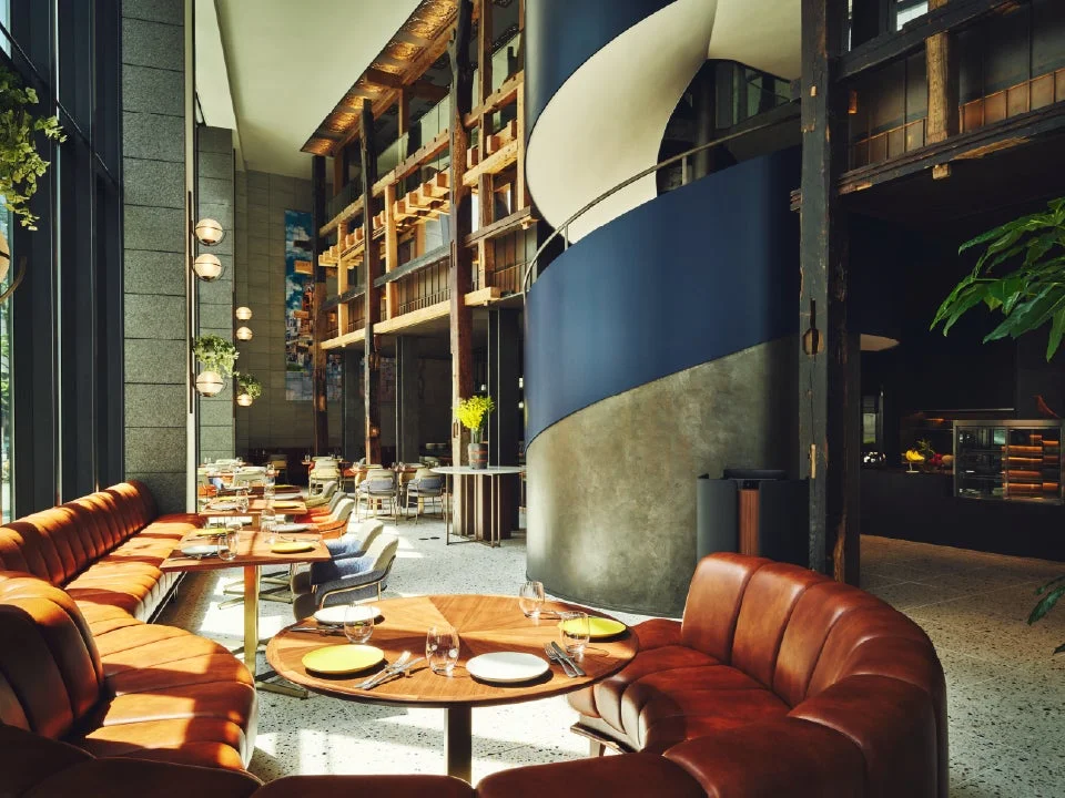 モダンでスタイリッシュなレストランまたはホテルラウンジの内部です。特徴的な螺旋階段、大きな窓、高い天井が印象的で、木材、コンクリート、革張りのソファが調和しています。テーブルには食器がセットされ、快適な食事ができる空間が広がっています。