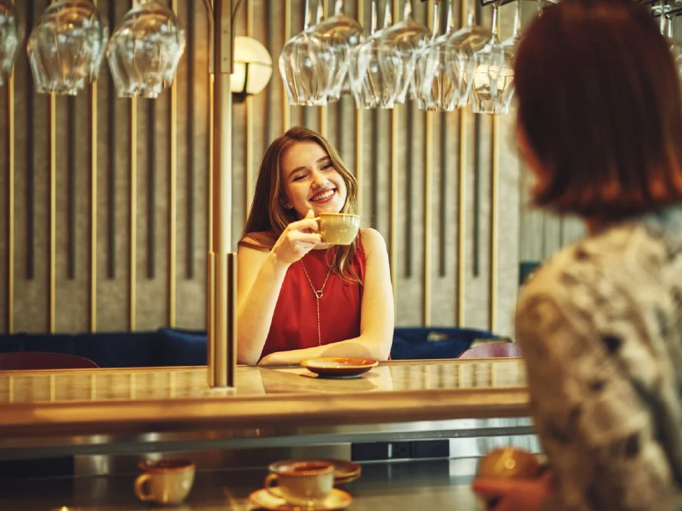 カフェのような場所で、赤い服を着た女性がコーヒーカップを手に笑顔で向かいの人と会話している様子。背景には多くのグラスが吊るされており、おしゃれな空間でリラックスした時間が流れています。