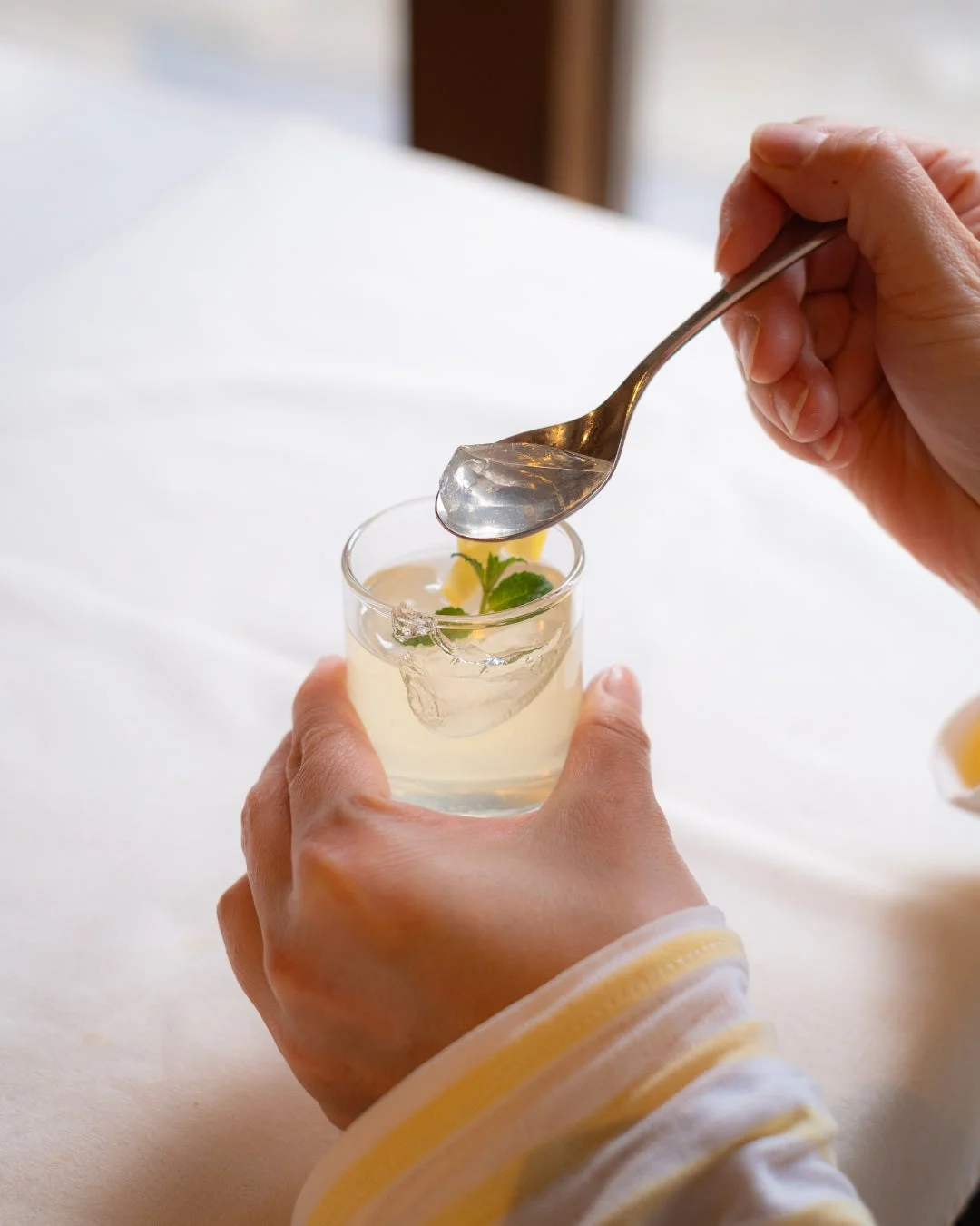 手がグラスを持ち、スプーンで透明なゼリーをすくっている様子。ミントとレモンが添えられた冷たいデザートドリンクで、爽やかな雰囲気が伝わります。