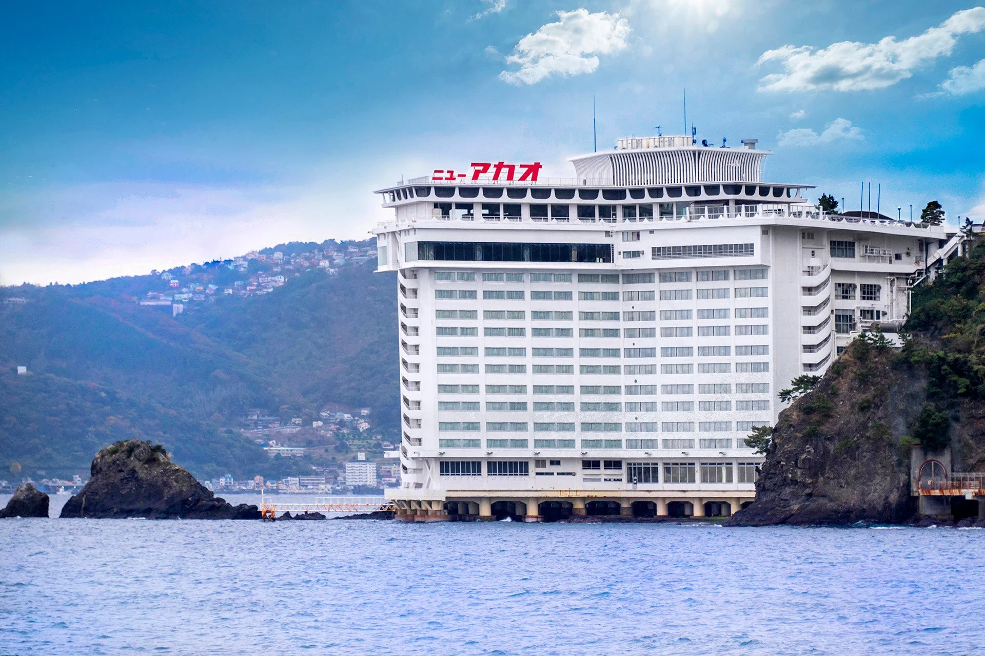 海に面して建つ巨大な白いホテル「ニューアカオ」の全景が写っています。背後には山々と住宅街が広がり、手前には岩場と青い海が広がっています。