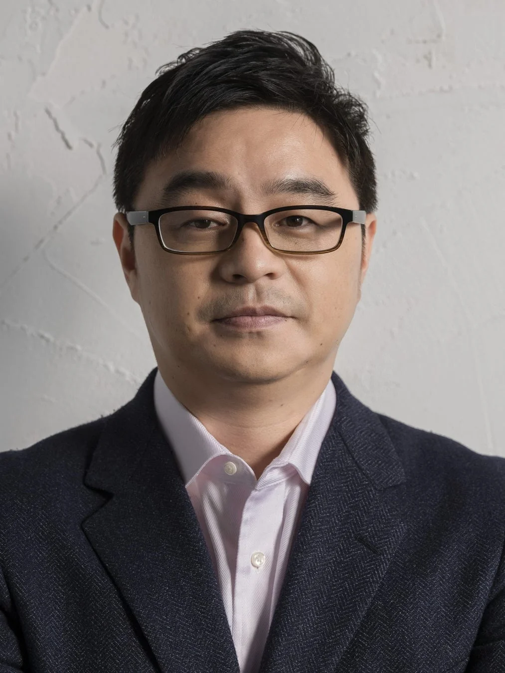 眼鏡をかけたアジア系の男性が、ピンクのシャツとダークなスーツジャケットを着用し、正面を向いている上半身のポートレート画像。