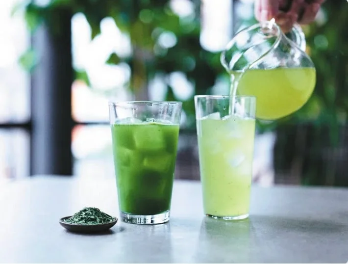 氷の入った二つのグラスに緑色の飲み物が注がれており、その傍らには抹茶のような緑の粉末が添えられています。背景には緑の植物がぼかされており、爽やかで健康的なドリンクのシーンを表現しています。