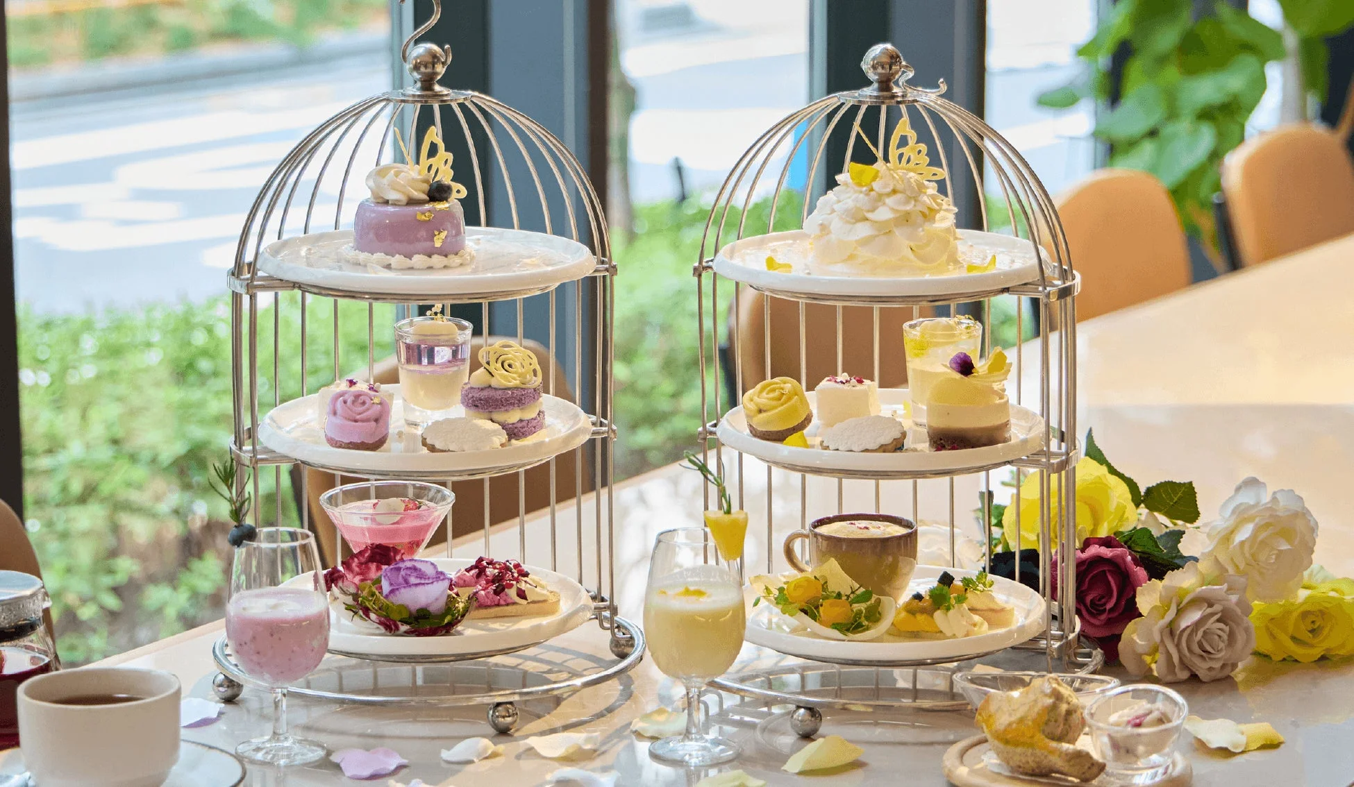 窓際で提供される鳥かご型のスタンドに並んだ、色とりどりの華やかなアフタヌーンティーセット。美しいデザートやドリンク、テーブルを飾る花々が優雅なティータイムを演出しています。