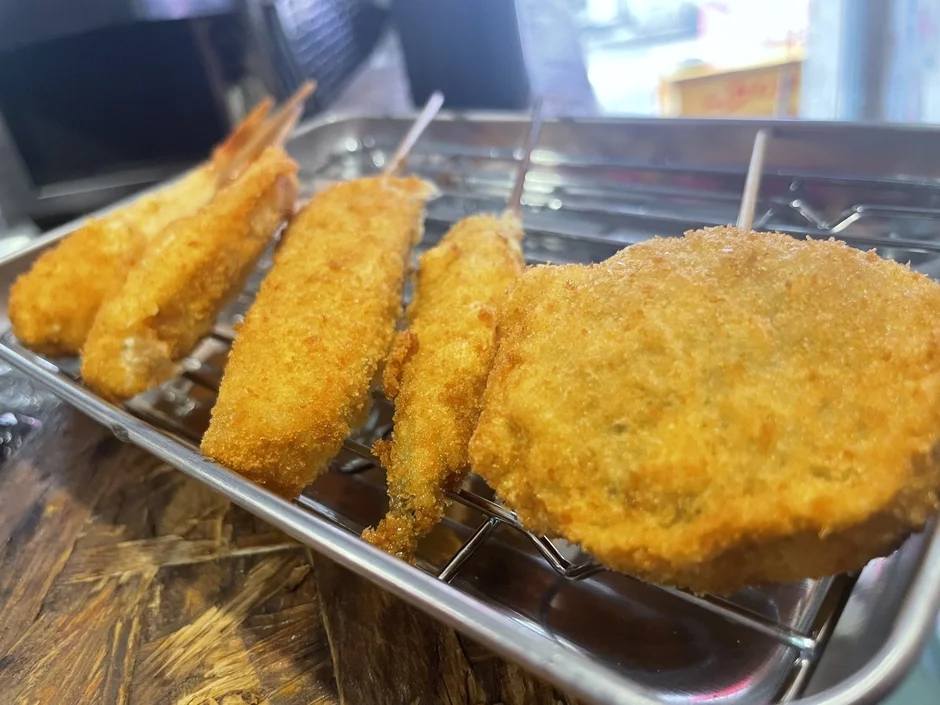 揚げたての串カツやエビフライなど、様々な揚げ物が金属製の網に乗せられて提供されている様子。きつね色に揚がった衣が食欲をそそります。