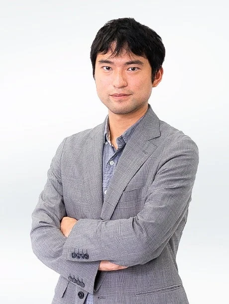 白い背景の前で、グレーのスーツと水色のシャツを着たアジア人男性が腕を組んで立っています。カメラをまっすぐ見つめており、真面目な印象を与えます。