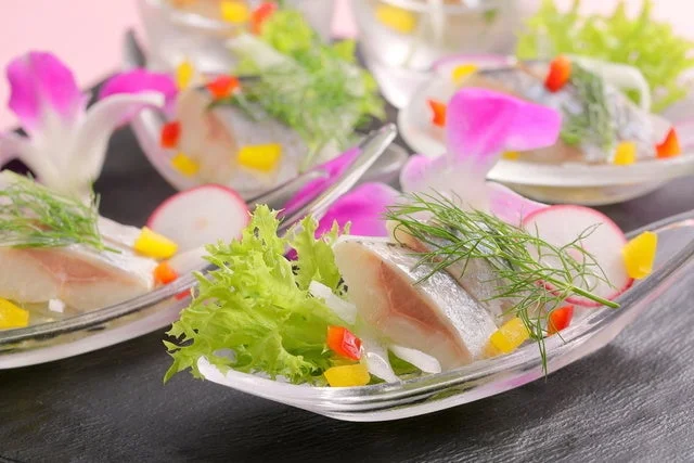透明な皿に盛り付けられた、魚と野菜の彩り豊かな前菜。ディル、ラディッシュ、パプリカが添えられ、ピンク色の装飾が華やかさを加えています。