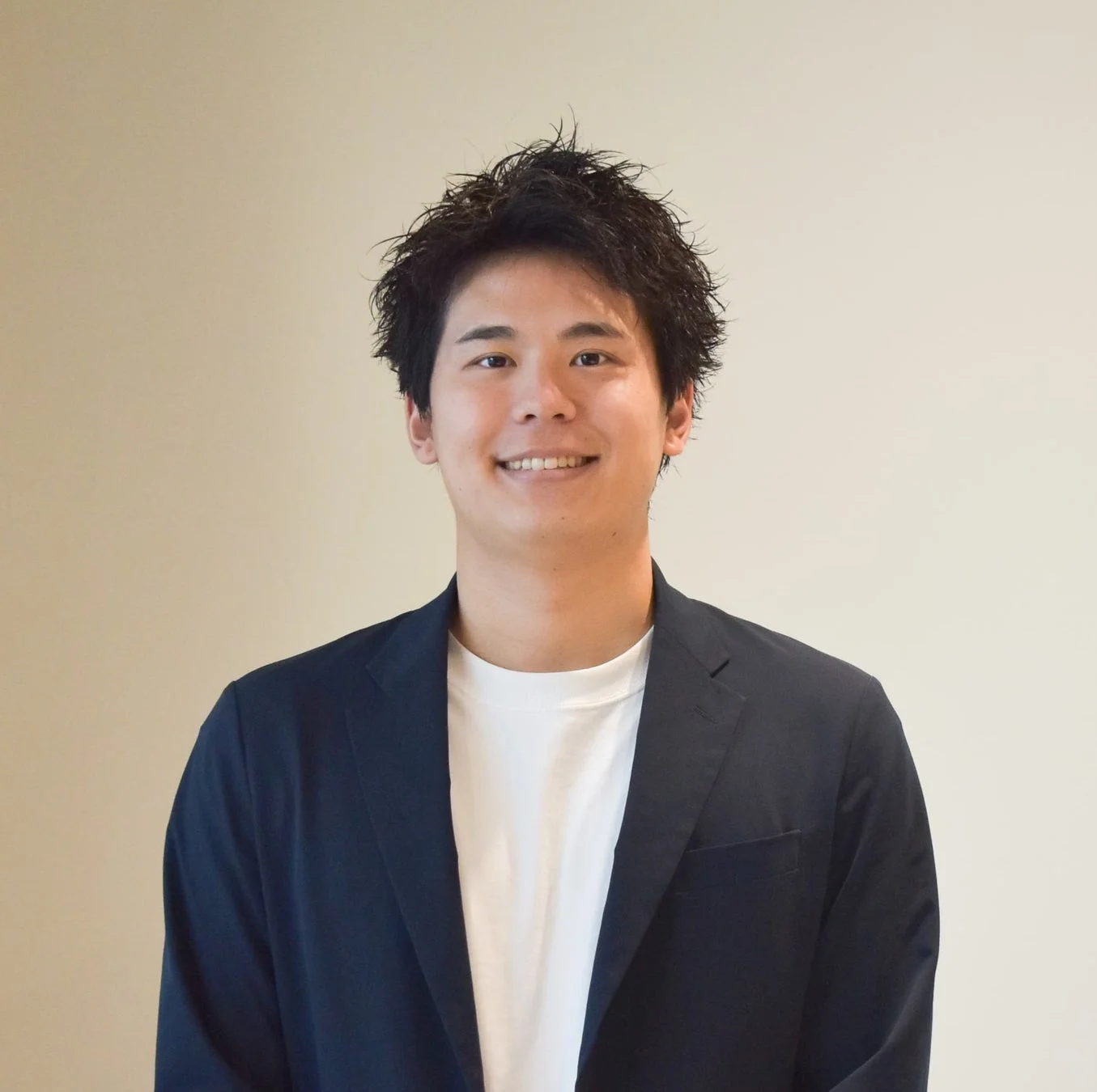 若い日本人男性が白いTシャツとネイビーのジャケットを着用し、笑顔で正面を見ているポートレート写真です。背景はシンプルなオフホワイトの壁です。