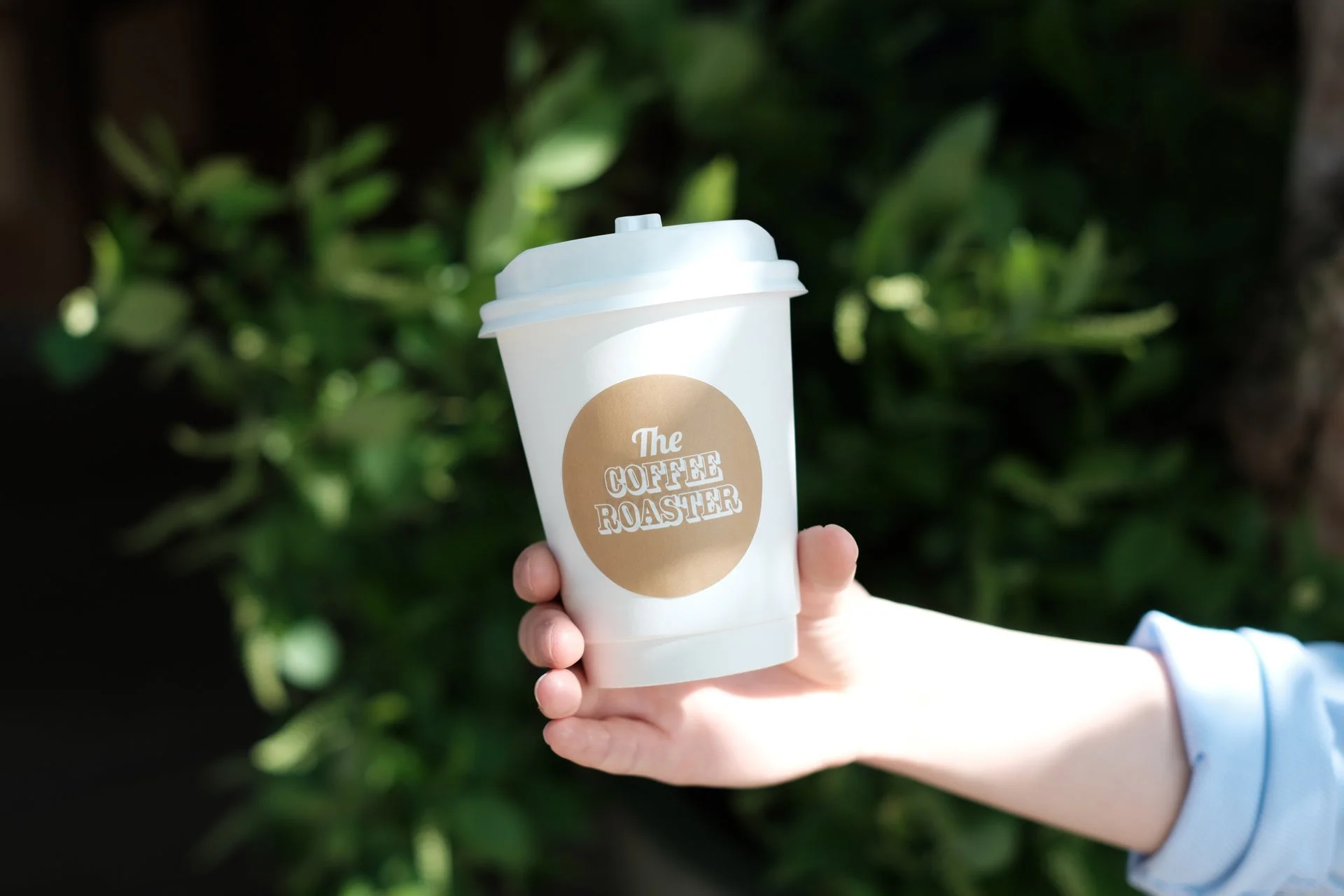 屋外で、手で「The COFFEE ROASTER」のロゴが入った白いコーヒーカップを持っている様子