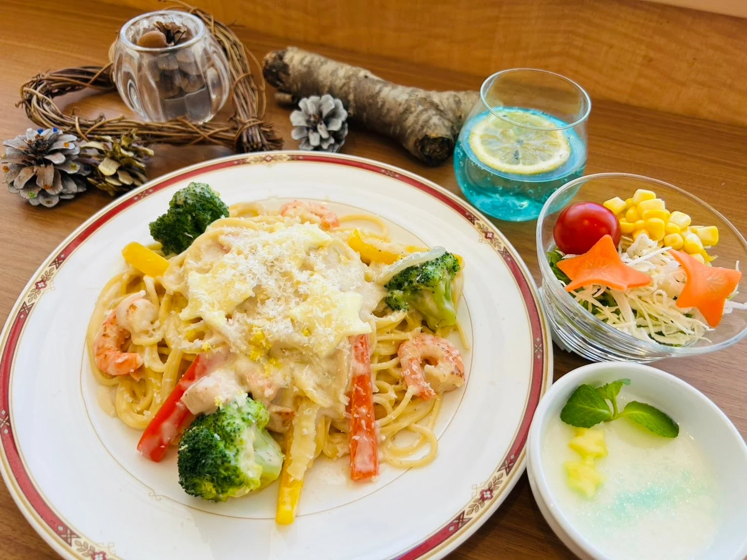 エビと彩り野菜がたっぷり入ったクリームパスタを中心に、サラダ、デザート、レモン入りドリンクが並べられた、カフェ風の食卓です。