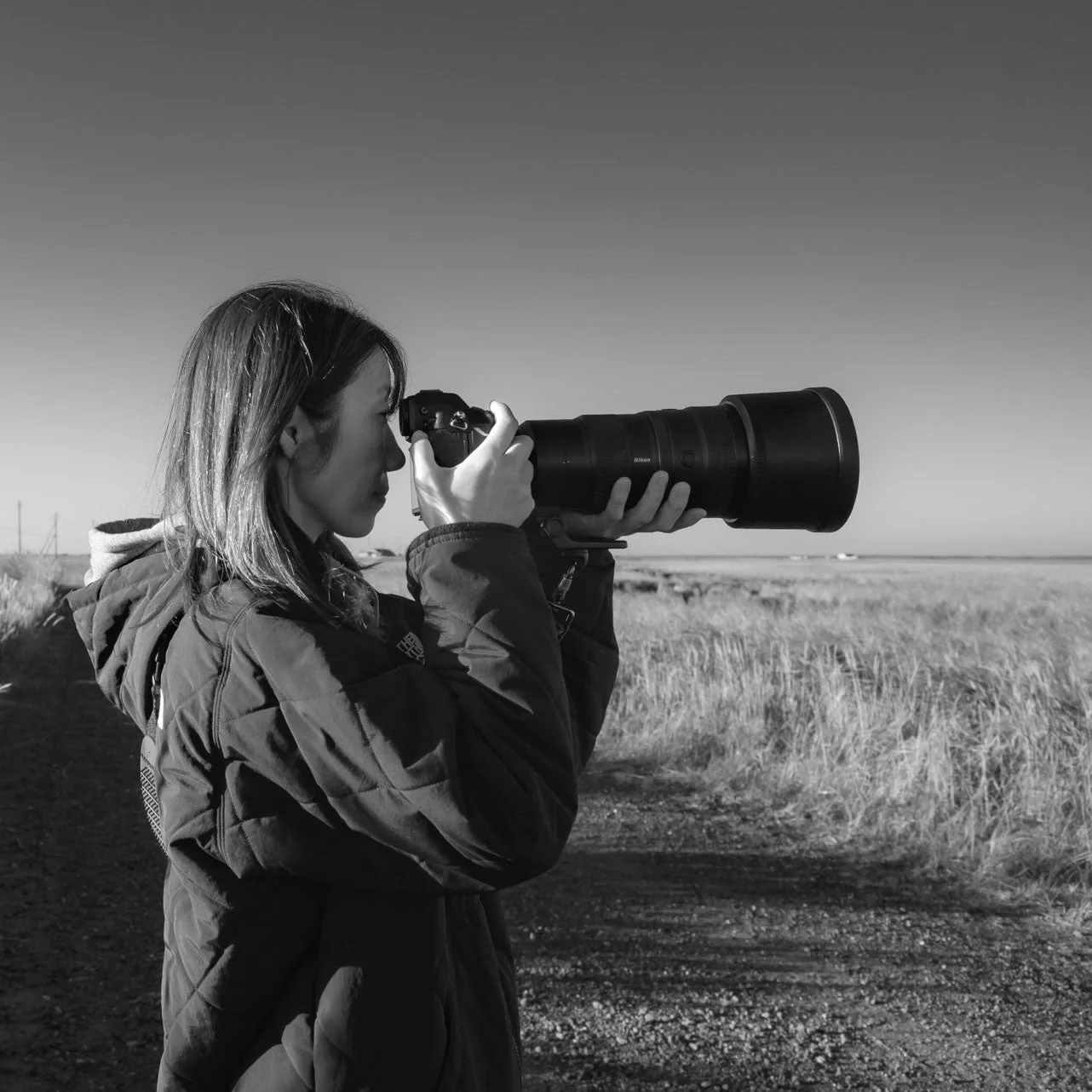 広大な屋外で、女性が望遠レンズ付きのカメラを構え、熱心に撮影している様子を捉えた白黒写真。