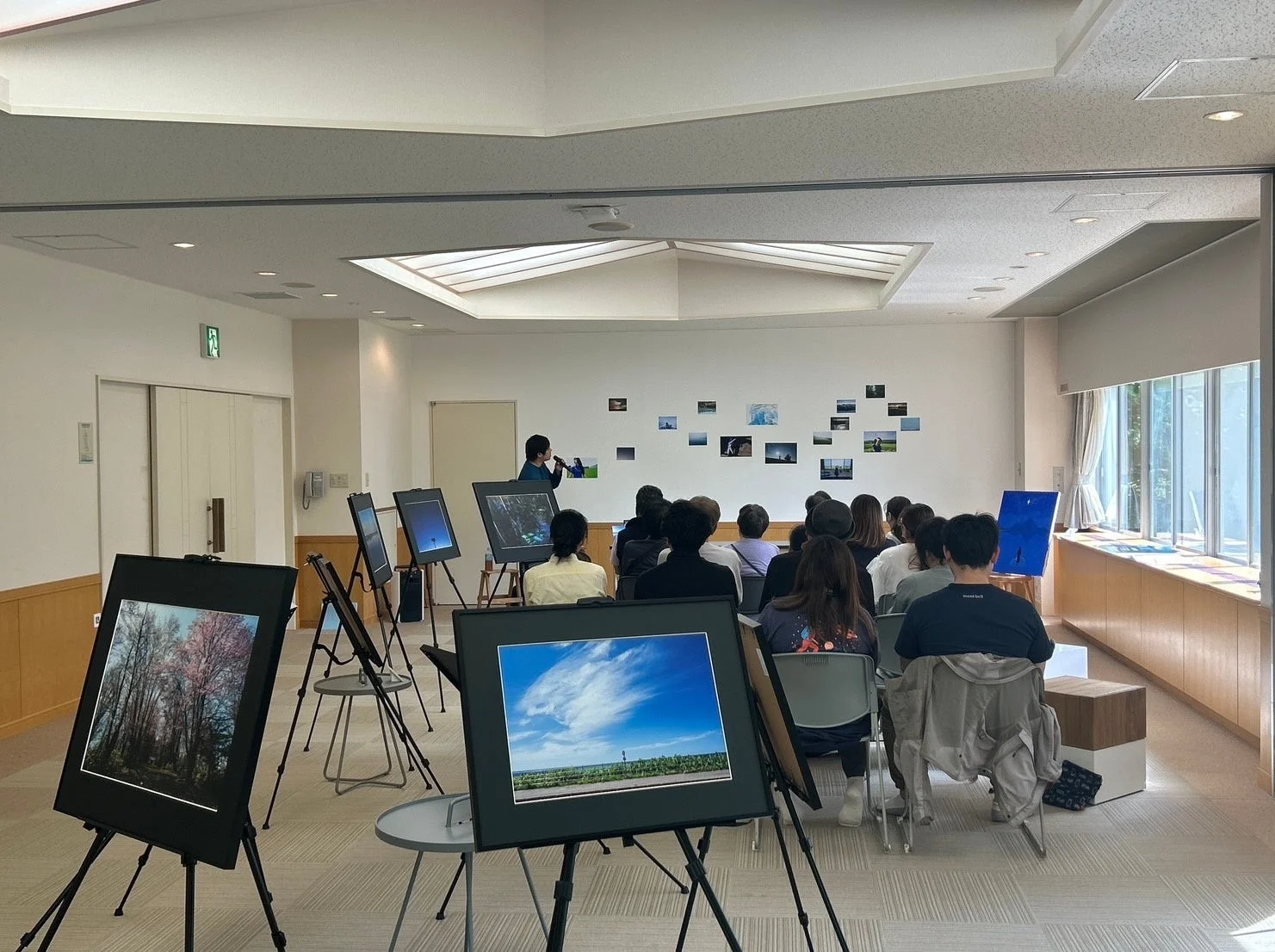 屋内会場で写真展が開催されており、講演者が聴衆にプレゼンテーションを行っている様子。会場には自然風景をテーマにした写真が多数展示され、参加者は熱心に鑑賞している。