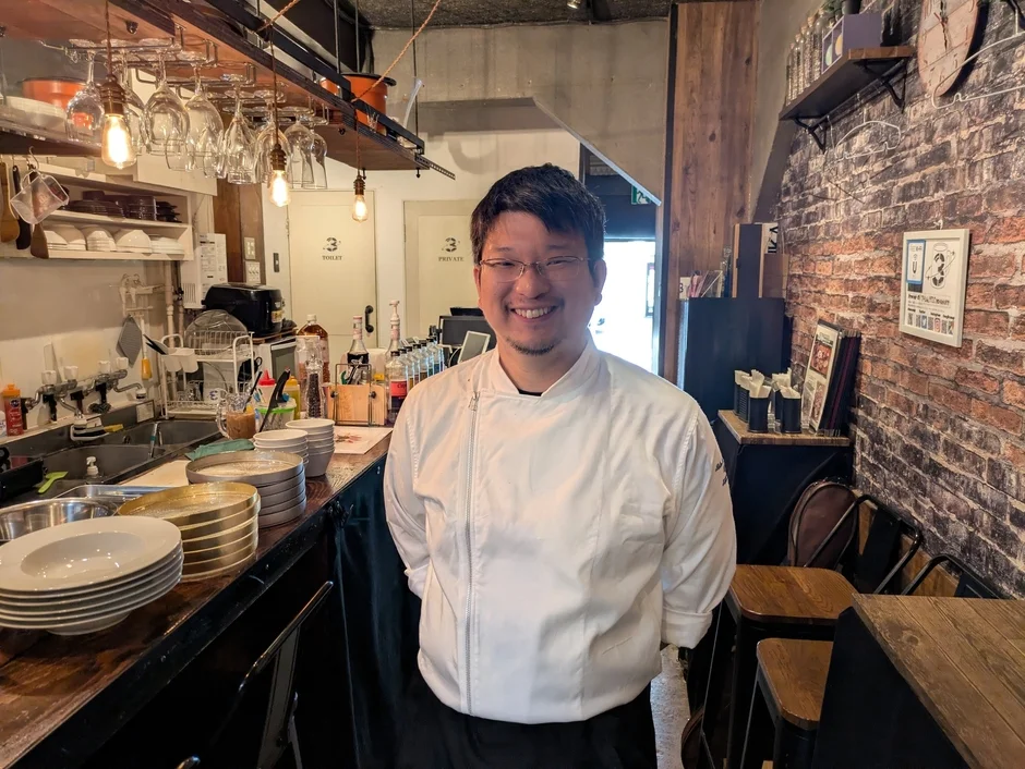 白いコック服を着た男性シェフが、レストランの厨房またはバーカウンターで笑顔を見せています。背景には調理器具や食器、レンガ壁が見え、活気ある飲食店の雰囲気が伝わります。