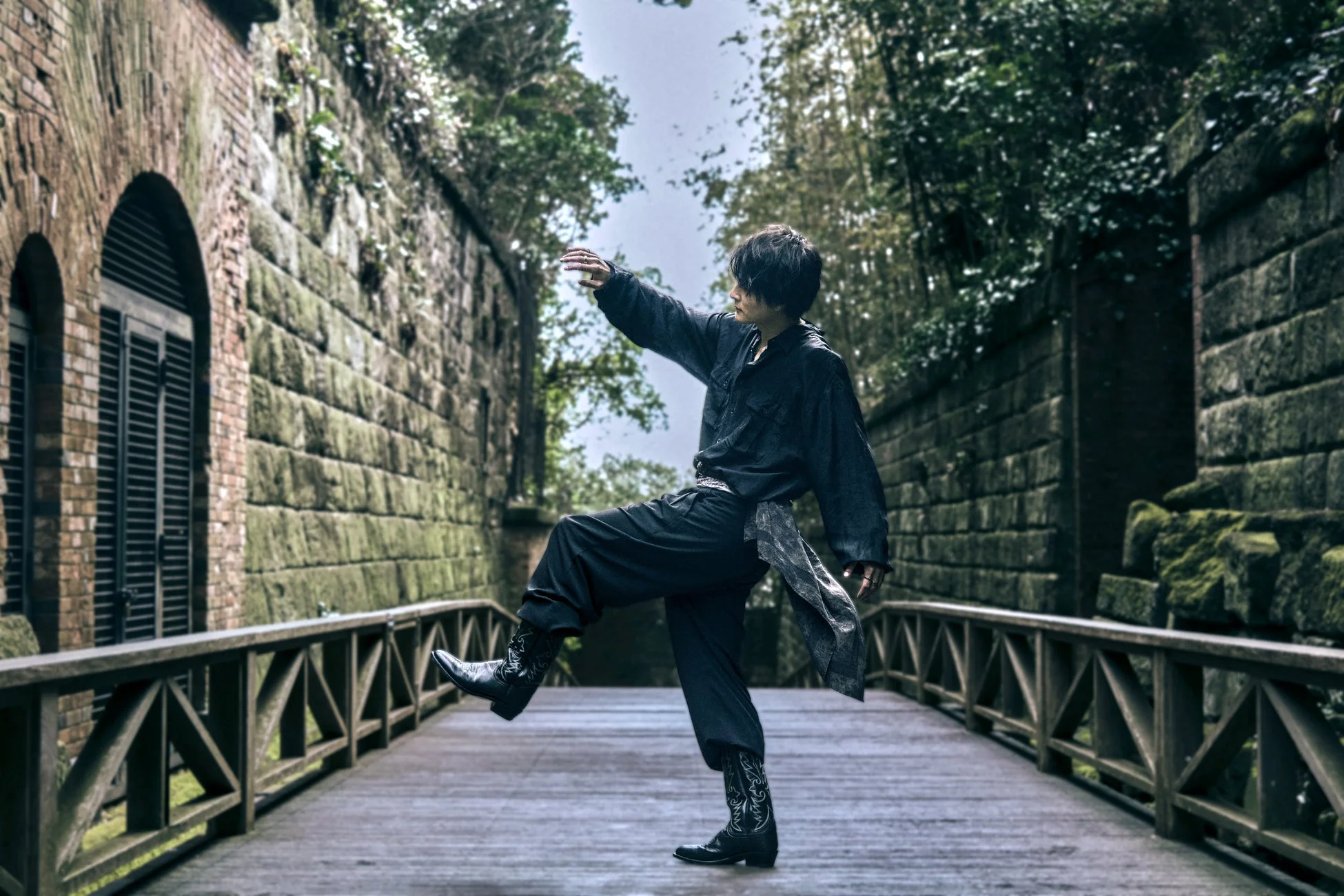 暗い服装とカウボーイブーツを身につけた男性が、石壁と木々に囲まれた木製の橋の上で片足を上げてポーズをとっている。神秘的な雰囲気の屋外ポートレート。