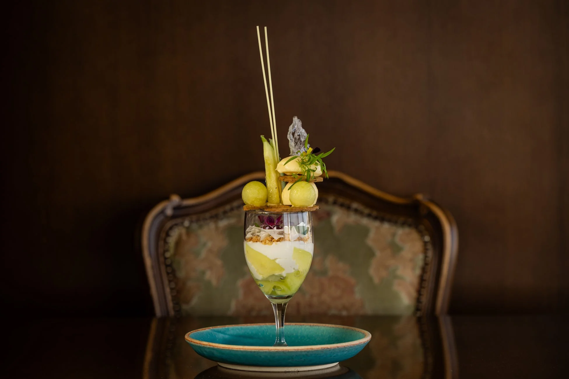 ワイングラスに美しく盛り付けられた、メロンやクリームを使った爽やかなグリーン系のパフェです。豪華なトッピングと優雅な背景が、特別なデザート体験を演出しています。