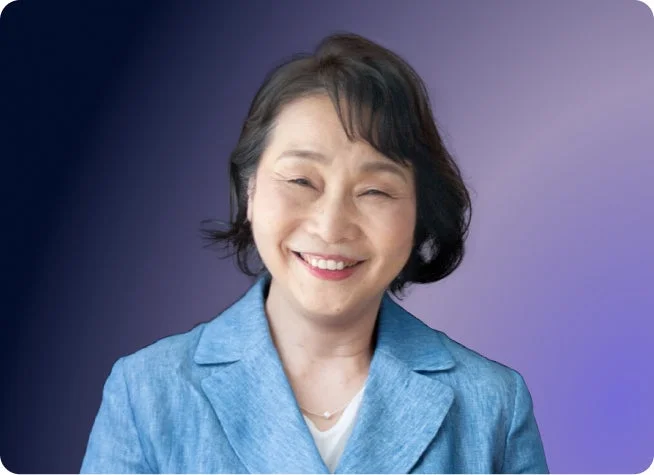 青いジャケットを着用したアジア系の女性が、紫色のグラデーション背景の前で優しい笑顔を見せているポートレート写真です。