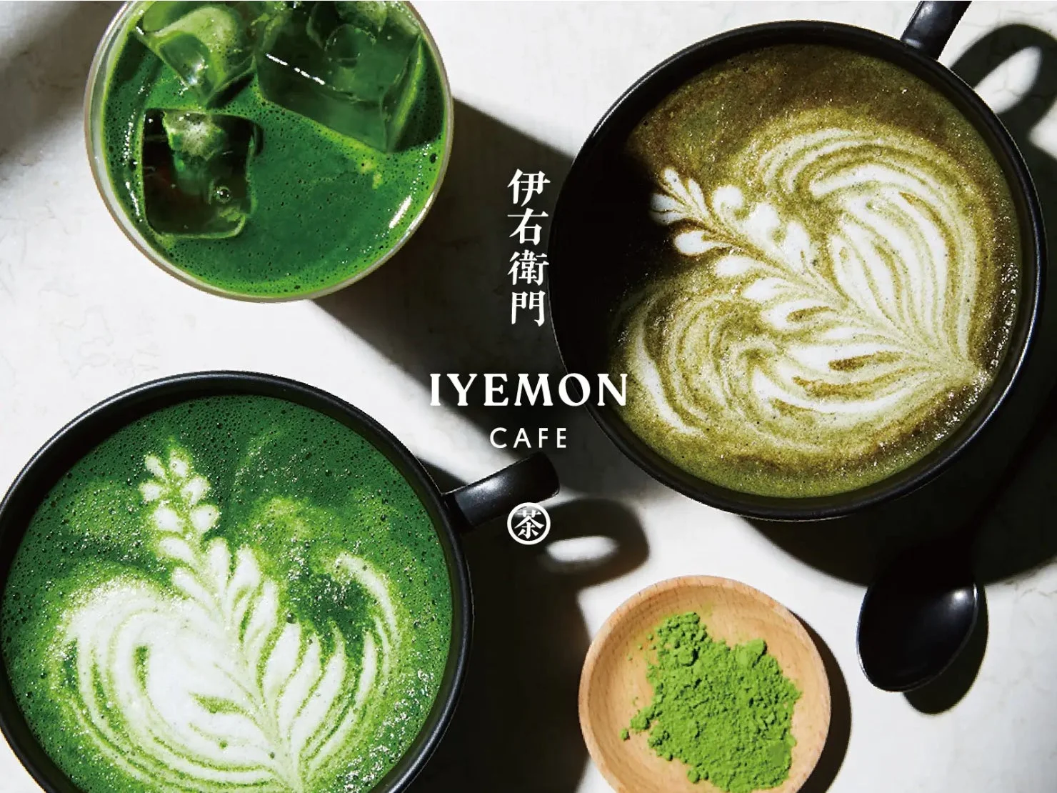 伊右衛門カフェの様々な抹茶ドリンクが写された画像です。美しいラテアートが施された抹茶ラテ2杯と、氷の入ったアイス抹茶、そして抹茶粉末が木製の器に入っています。鮮やかな緑色が印象的な、魅力的なカフェの風景です。